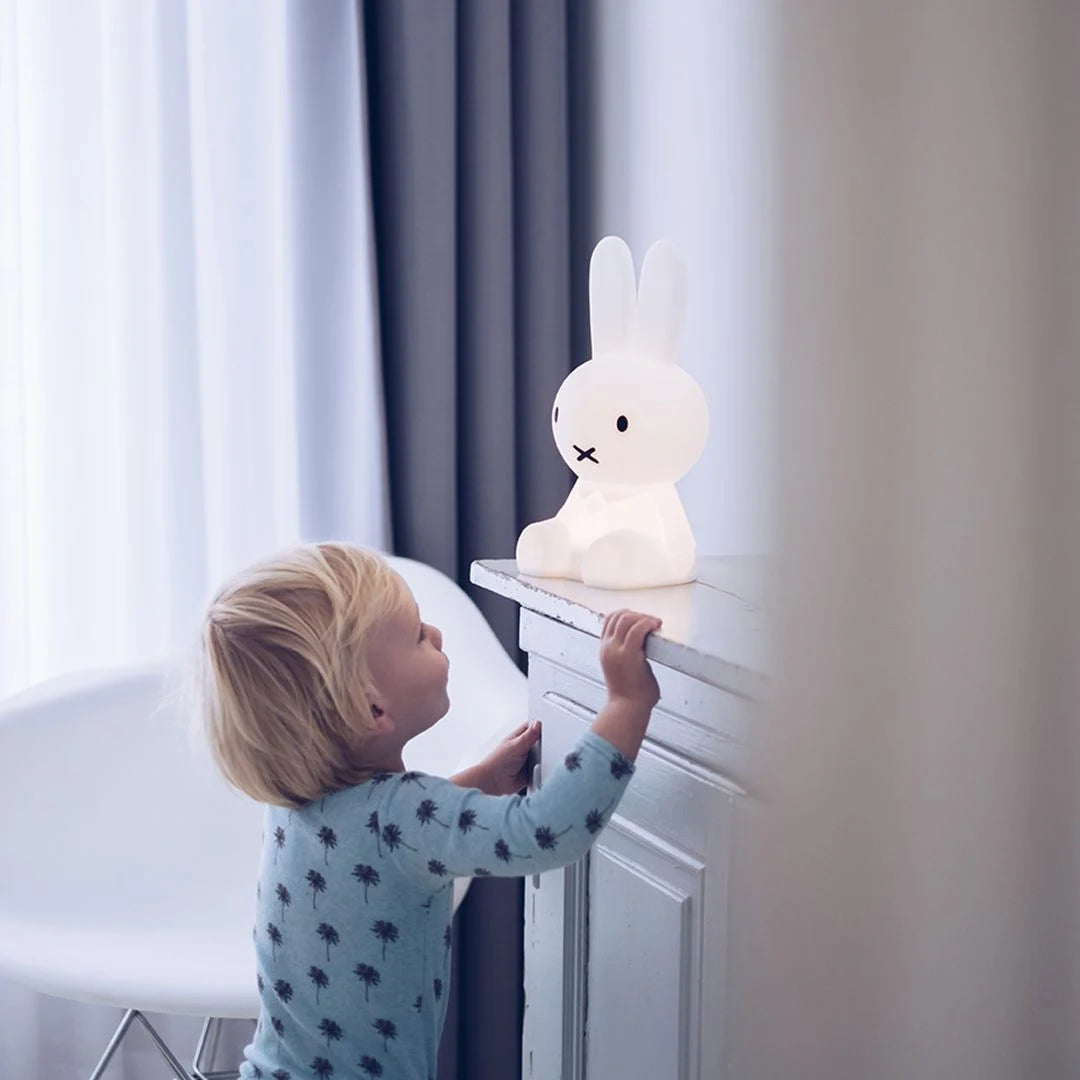A young child wearing blue pajamas with a palm tree pattern looks up at the glowing Miffy First Light Lamp placed on a white mantelpiece.