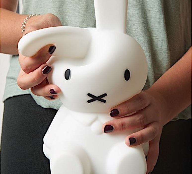 A white Miffy First Light Lamp shaped like a rabbit with long ears and a simple black-lined face. A woman's hands holding the Miffy folding one ear