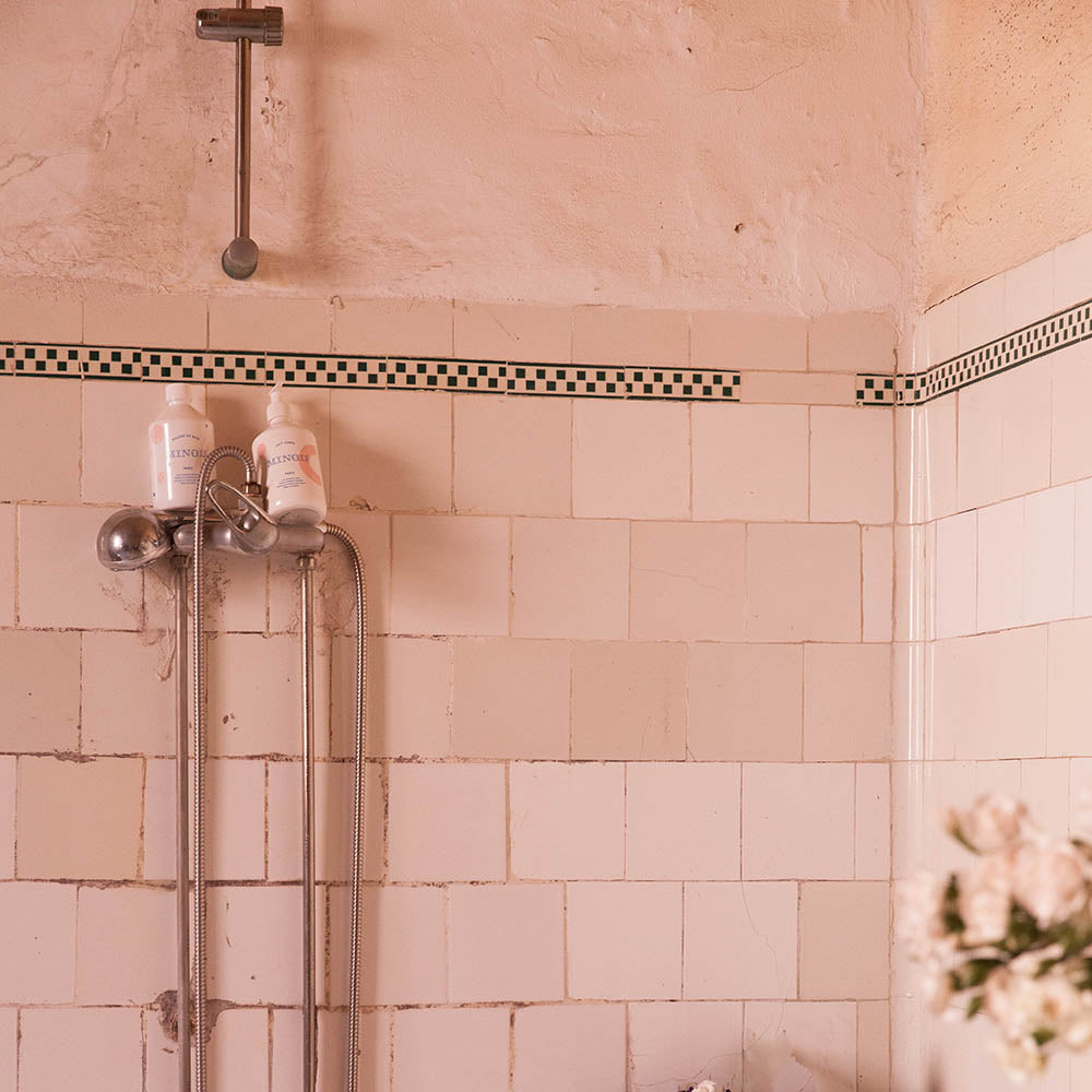Two minois products, sit on top of a shower head inside a shower room that has pink tiles and a black checkered pattern above it. Above the tiling of the shower, the room is pink and there is some silver pipe work. There are also some pink roses in the image at the right hand side of the shower.
