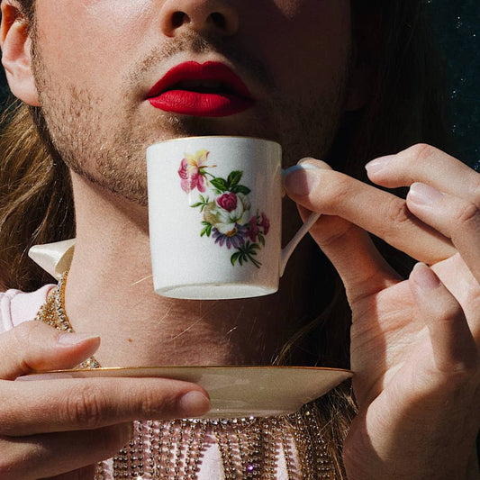 An image of somebody with red lipstick and a gold necklace on holding a small cup with pink yellow purple and green flowers on up to their lips whilst holding a saucer in the other hand.