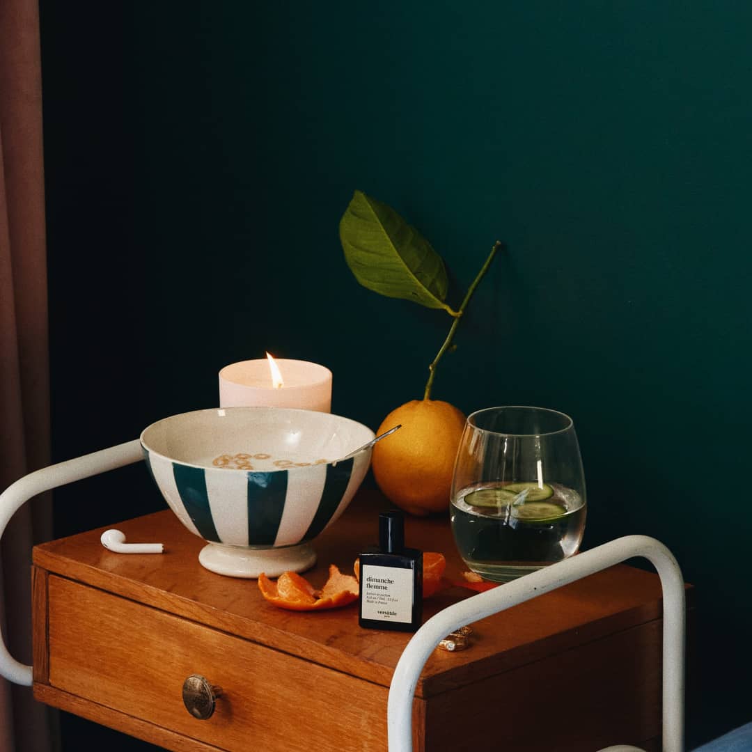 An image of a brown beside table that has an orange with a long green stem on it, a white bowl with green stripes containing milk and cereal, a clear glass with clear liquid and cucumbers in it, a white candle burning, an AirPod and a bottle of dimanche flemme perfume.