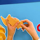 Hands assembling a yellow paper craft on a blue background with a red mug and spoon.