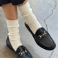 Black loafers with gold buckles worn with white socks on a concrete floor.