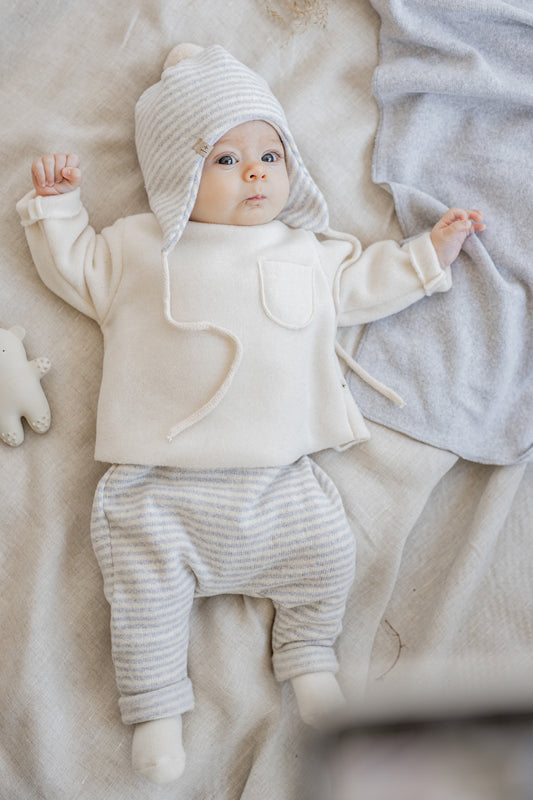 on a linen beige bed sheet next to a red blanket. a baby sprawled with arms and legs outstretched wearing a striped grey and white bonnet and harem bottoms. also wearing a white fleece jumper with a little patch pocket on the chest