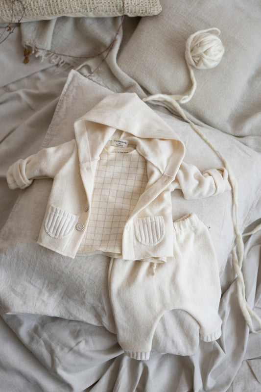 on a grey linen bed sheet is a hooded cardigan in white open with arms spears outward with two little ribb pockets wither side of the cardigan front. and a white harem pant with ribb cuffs