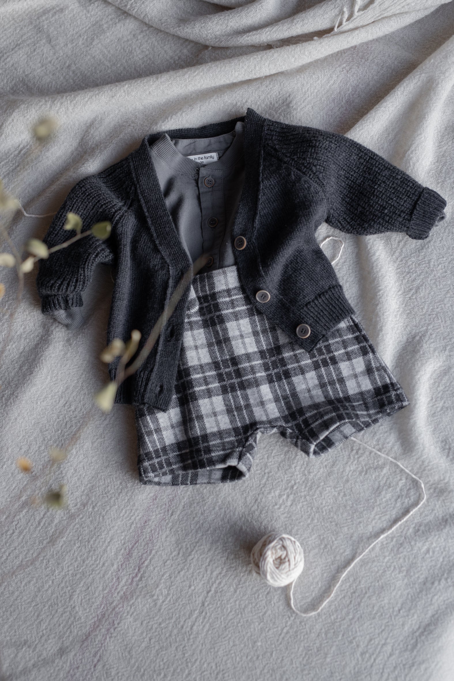 on a linen bed sheet a charcoal grey cardigan open over a grandpa colar shirt with a pair of tartan grey shorts