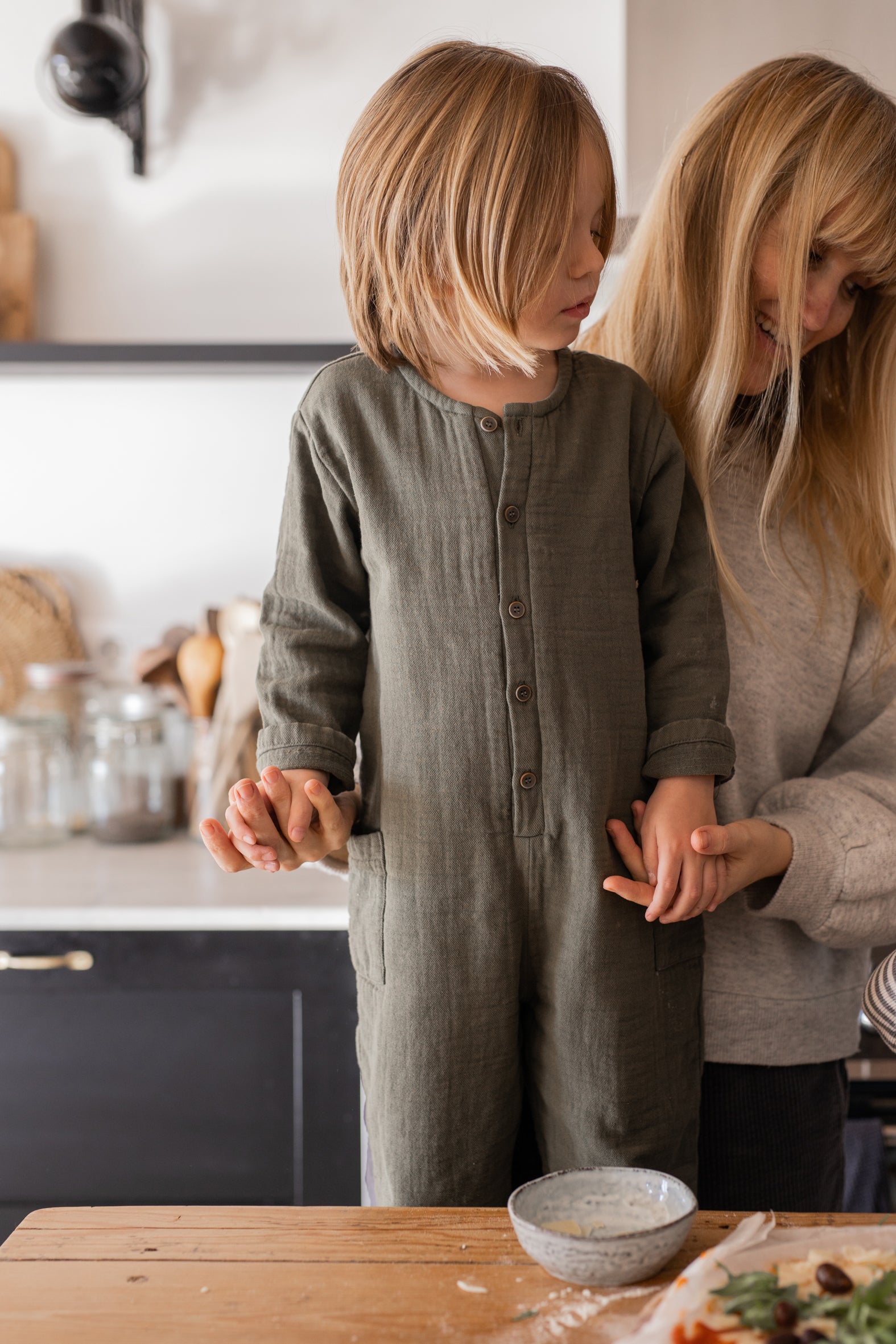 a child in the kitchen being held by its mother. wearing the green jumpsuit, having the sleeves cuffed at the wrists