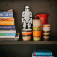 A set of HKliving 70's ceramic coffee mugs sits on a dark shelf surrounded by a pile of books and a Star Wars Stormtrooper figure