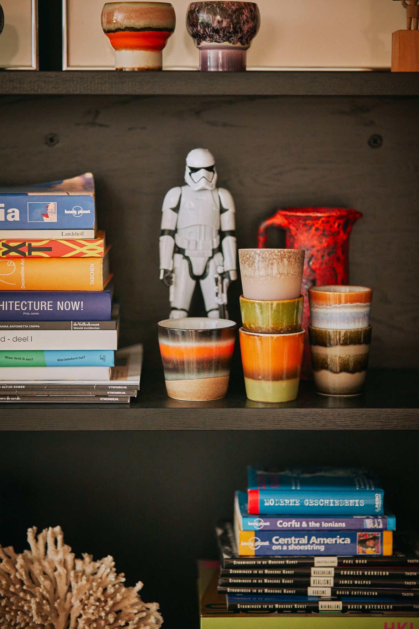 A set of HKliving 70's ceramic coffee mugs sits on a dark shelf surrounded by a pile of books and a Star Wars Stormtrooper figure