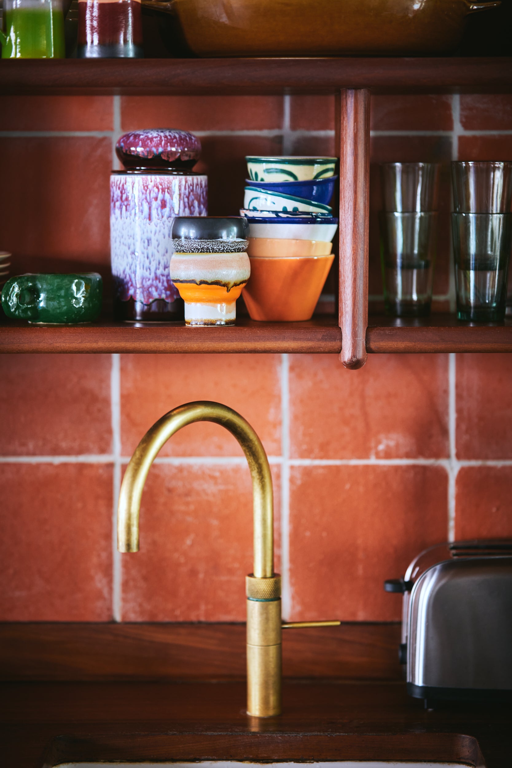 A collection of HKliving 70's ceramics sits on a shelf in a brown tiled kitchen in front of a gold curved tap