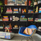 A stylish living space with black painted walls. A  colourful rug lies on the floor with a blue Eames rocking chair with the pink and yellow diagonal striped cushion on it. In the background are dark shelves on which various books, plants, pictures, and HKliving 70's ceramics are stacked.