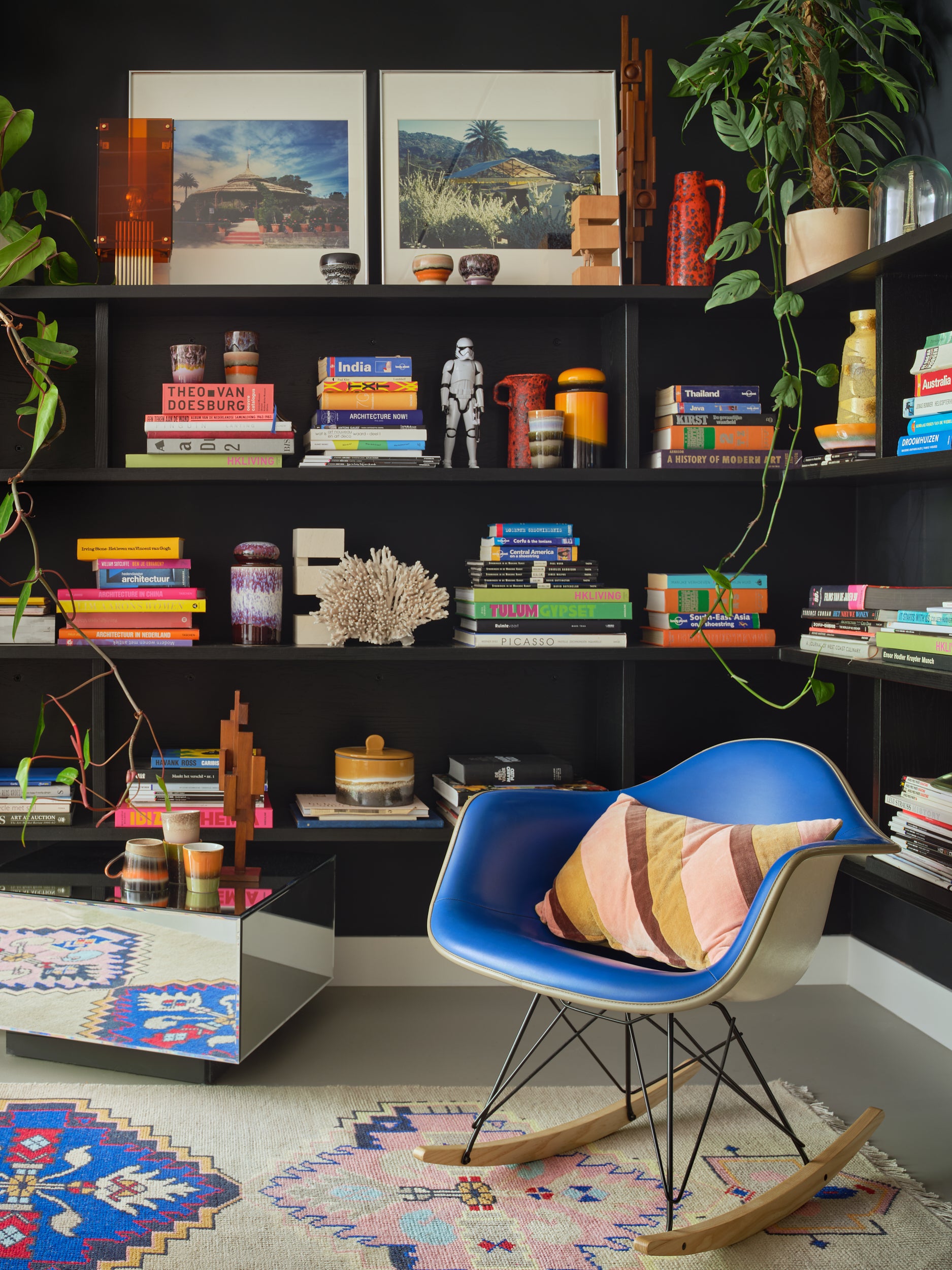 A stylish living space with black painted walls. A  colourful rug lies on the floor with a blue Eames rocking chair with the pink and yellow diagonal striped cushion on it. In the background are dark shelves on which various books, plants, pictures, and HKliving 70's ceramics are stacked.