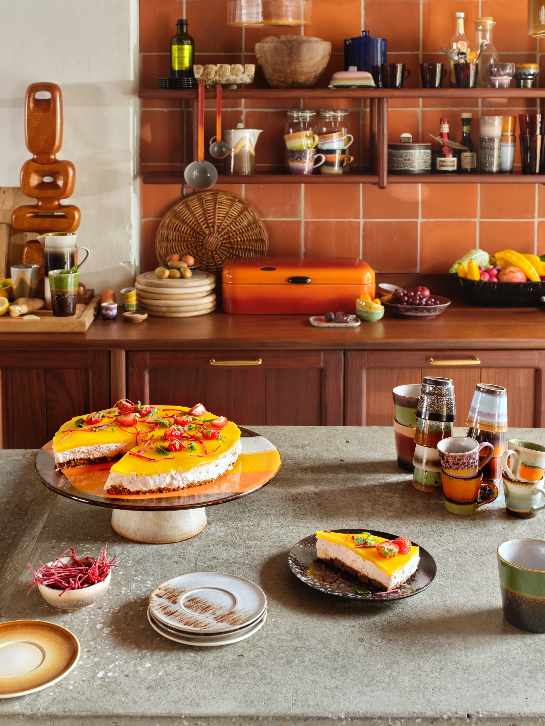 Here is a kitchen layout with hkliving mugs spread across the grey marble counter top and wooden shelves in the background. Next to a cake on a cake stand and other bows of snack displayed amongst the various mug collections. In the background there are stacked plates next to an organ be tool box and a bowl of fruit in front of the terracotta tiled wall