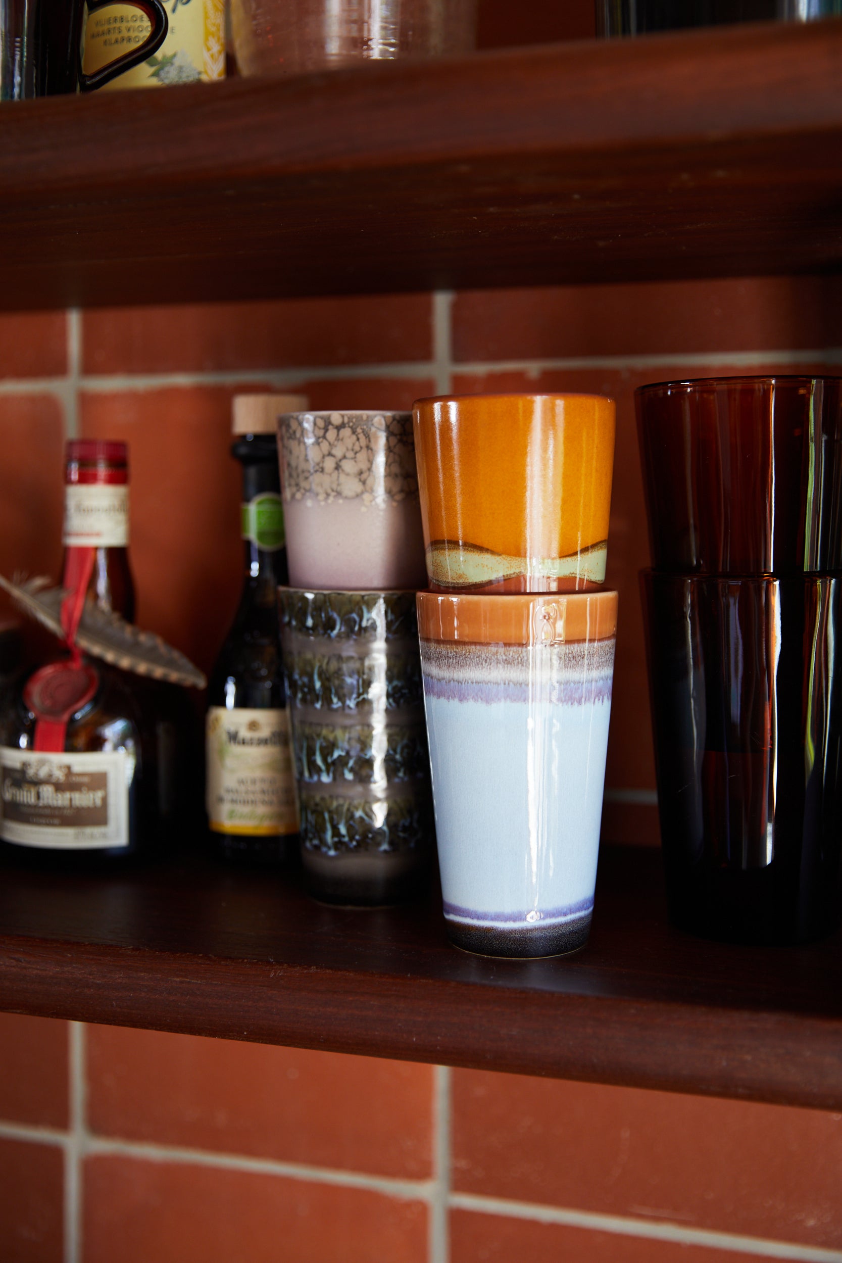 4 Hk living latte mugs in their different colours stacked 2 by 2 on a wooden shelf with two condiments and to brown glasses, in front of a terracotta tiled wall