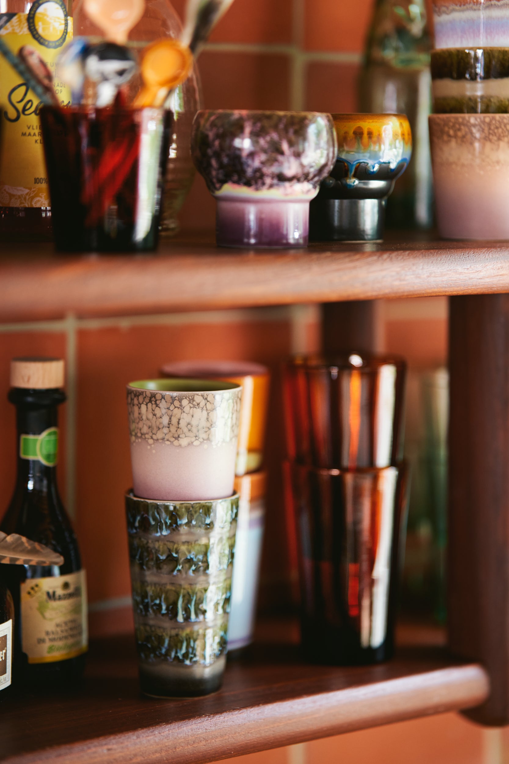 A variety of Hk living mugs on two open wooden shelves with different glasses and condiments displayed together. All in front of terracotta tiles on the wall