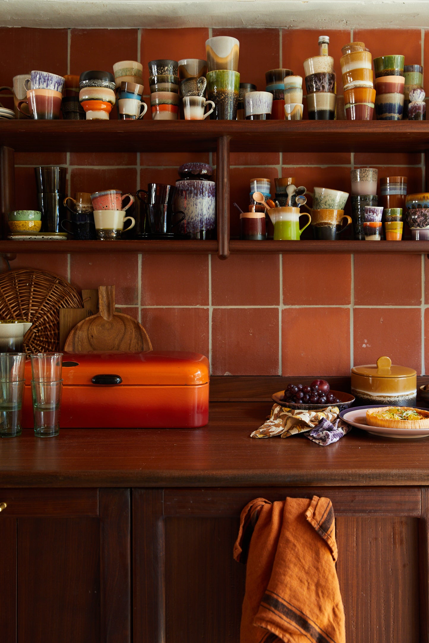 There are wooden shelves filed to the edges with a variety of Hk living mugs and bowls. In a kitchen setting with a large wooden counter top, that has an orange tool box on top with two plates of snacks and a Hk living pot. The background is has terracotta tiles all over the wall.