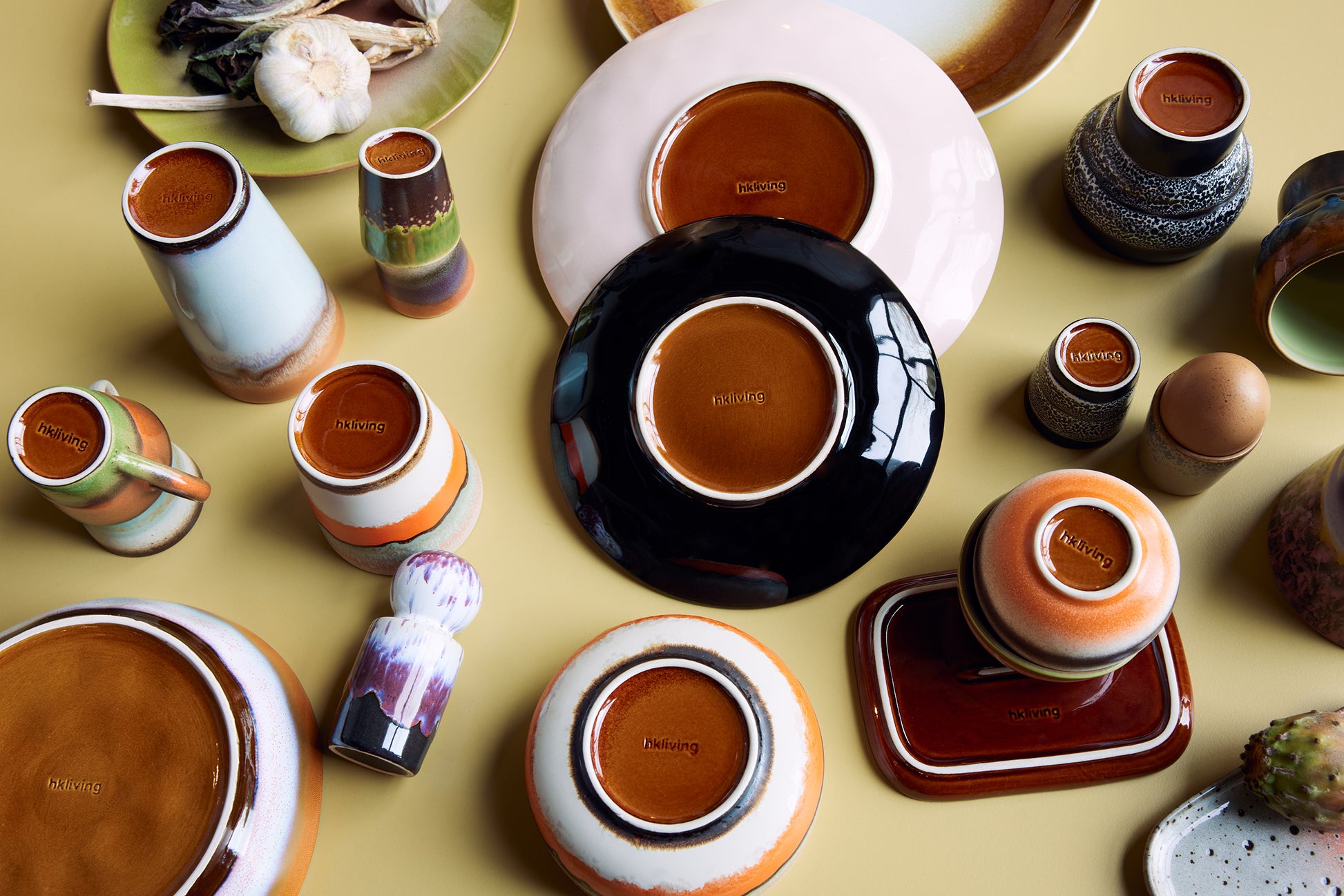 A flat lay of a variety of Hkliving plates, bowls and mugs. All turned over showing the embossed logo on their base in a cohesive brown colour. 