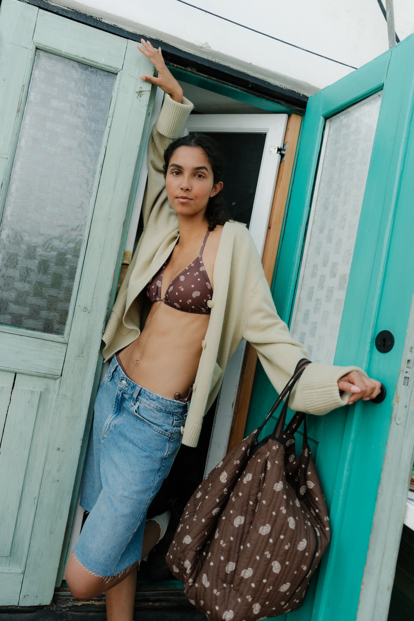 Woman in a bikini top and shorts standing in front of a turquoise door with a polka dot bag.