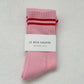 A pair of Le bon shoppe amour pink boyfriend socks which are pink in colour with red stripes at the top. The wrapping is white in colour with “Le Bon shoppe” and “boyfriend socks” written in black colour. The socks are kept against a white beige background 