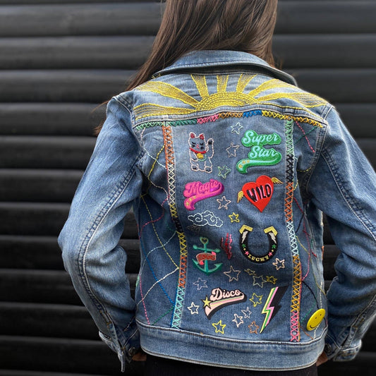 An image depicting a woman wearing a blue denim jacket. The jacket has a lot of colourful iron on patches. The background is grey. 