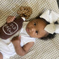 Baby lying on a patterned surface holding a knitted 'OLD BREW' bottle toy.