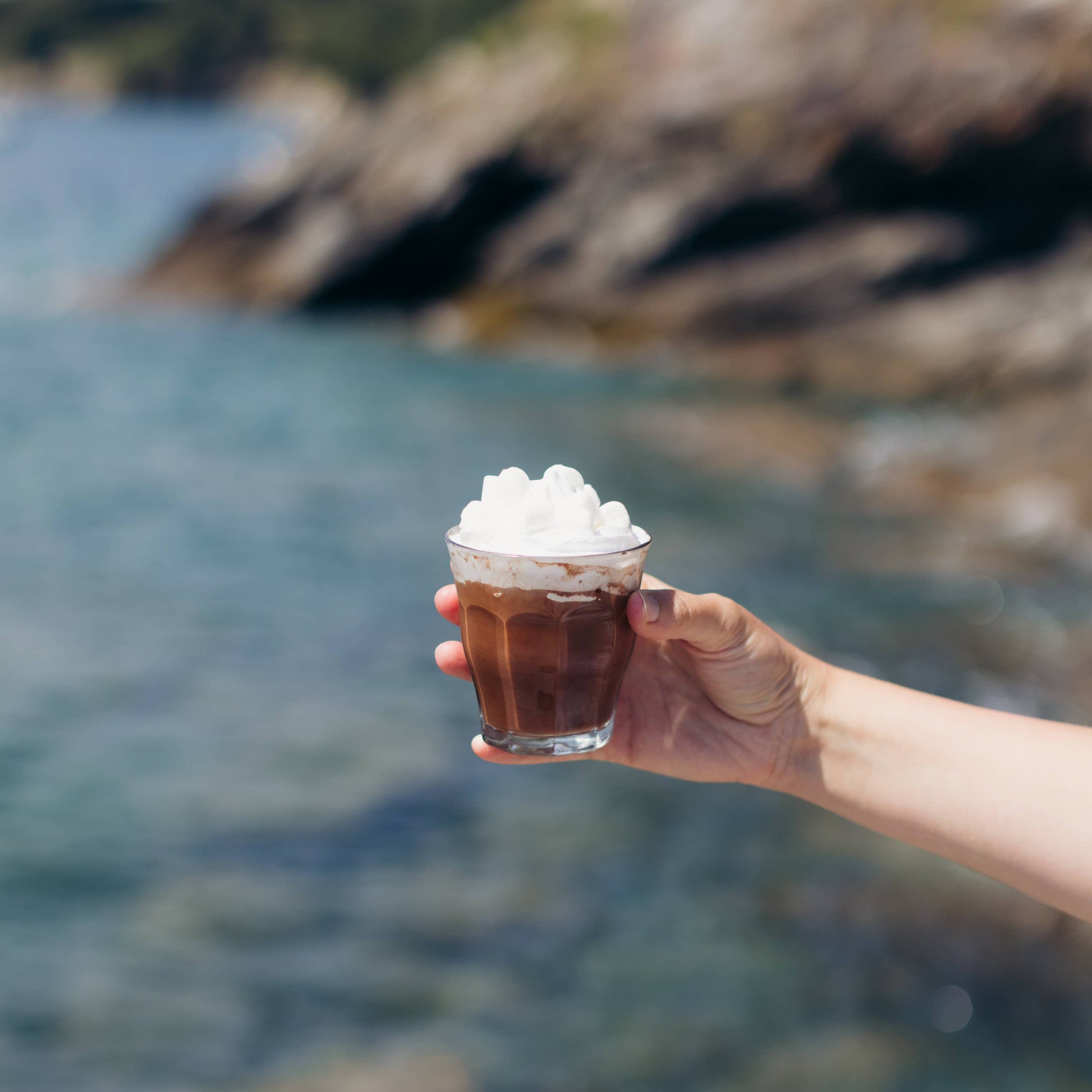 Hand holding a glass of chocolate milkshake with whipped cream against a blurred natural background