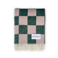 A folded Maison Deux blanket with a green and pink checkerboard pattern and fringes on the short sides, displaying the brand name on a white label.