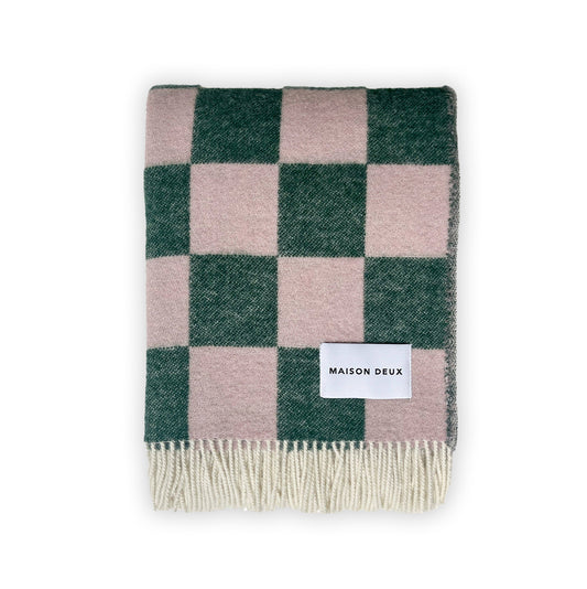 A folded Maison Deux blanket with a green and pink checkerboard pattern and fringes on the short sides, displaying the brand name on a white label.