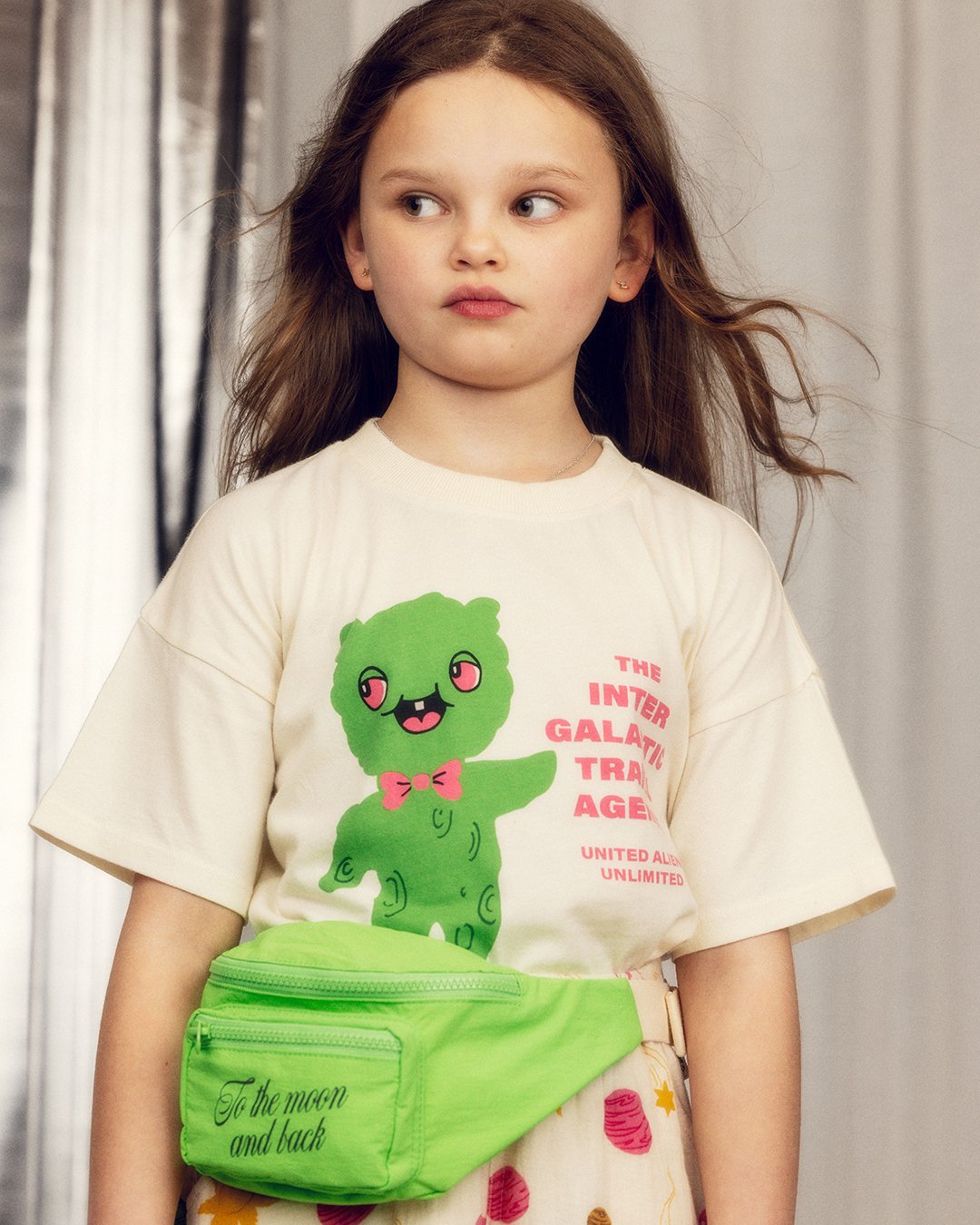 Young girl wearing a t-shirt with a green character and text, holding a green bag.