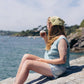Person sitting by a body of water holding a cup, wearing a light blue sleeveless shirt and denim shorts.