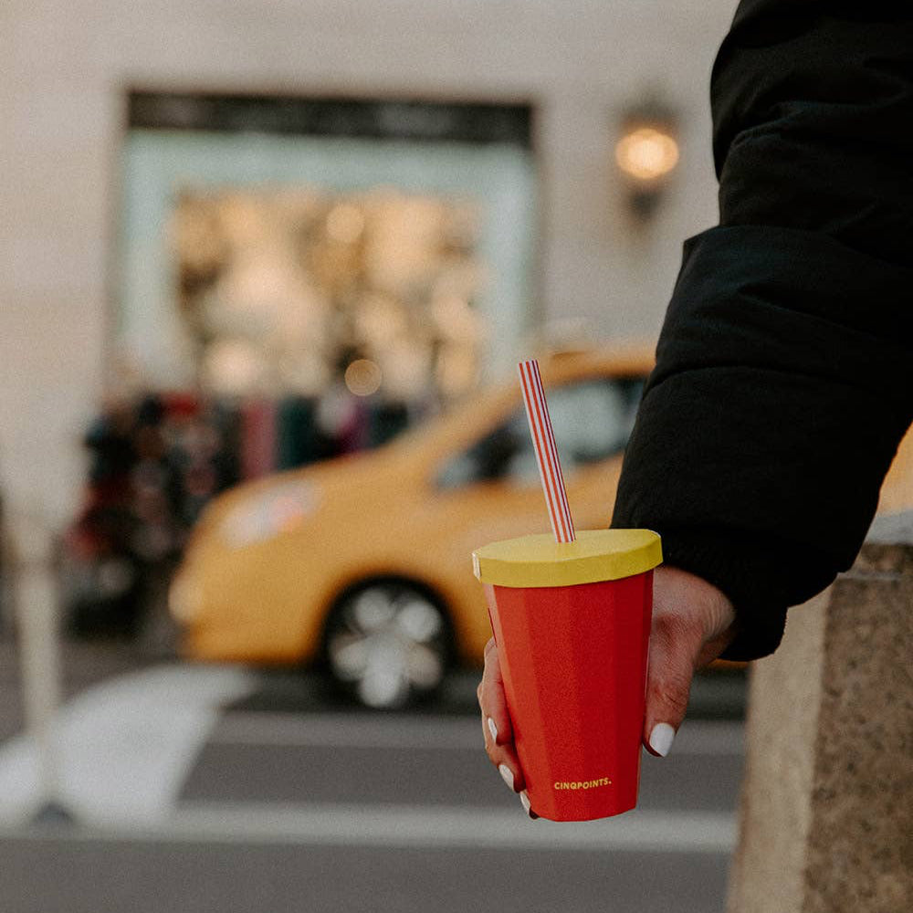 Person holding a red cup with a yellow lid and straw in an urban setting