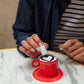 Person making a heart-shaped design in coffee with a red cup on a marble table.