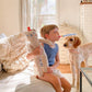 Child holding a teddy bear and blowing into a dog's nose in a cozy room.