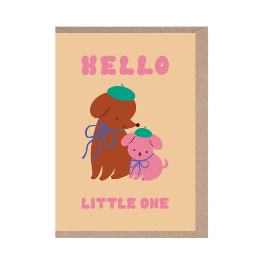 A greeting card with an illustration of two cartoon dogs wearing berets, one brown and one pink, with the text 'HELLO LITTLE ONE' above. The card is set against a beige background and has a recycled brown kraft envelope.