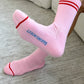 A pair of Le bon shoppe amour pink boyfriend socks which are pink in colour with red stripes at the top and near the toes are worn by a person. It says Le Bon shoppe on the sole of the socks in blue colour. The floor is wooden and is beige with brown lines of detail. 