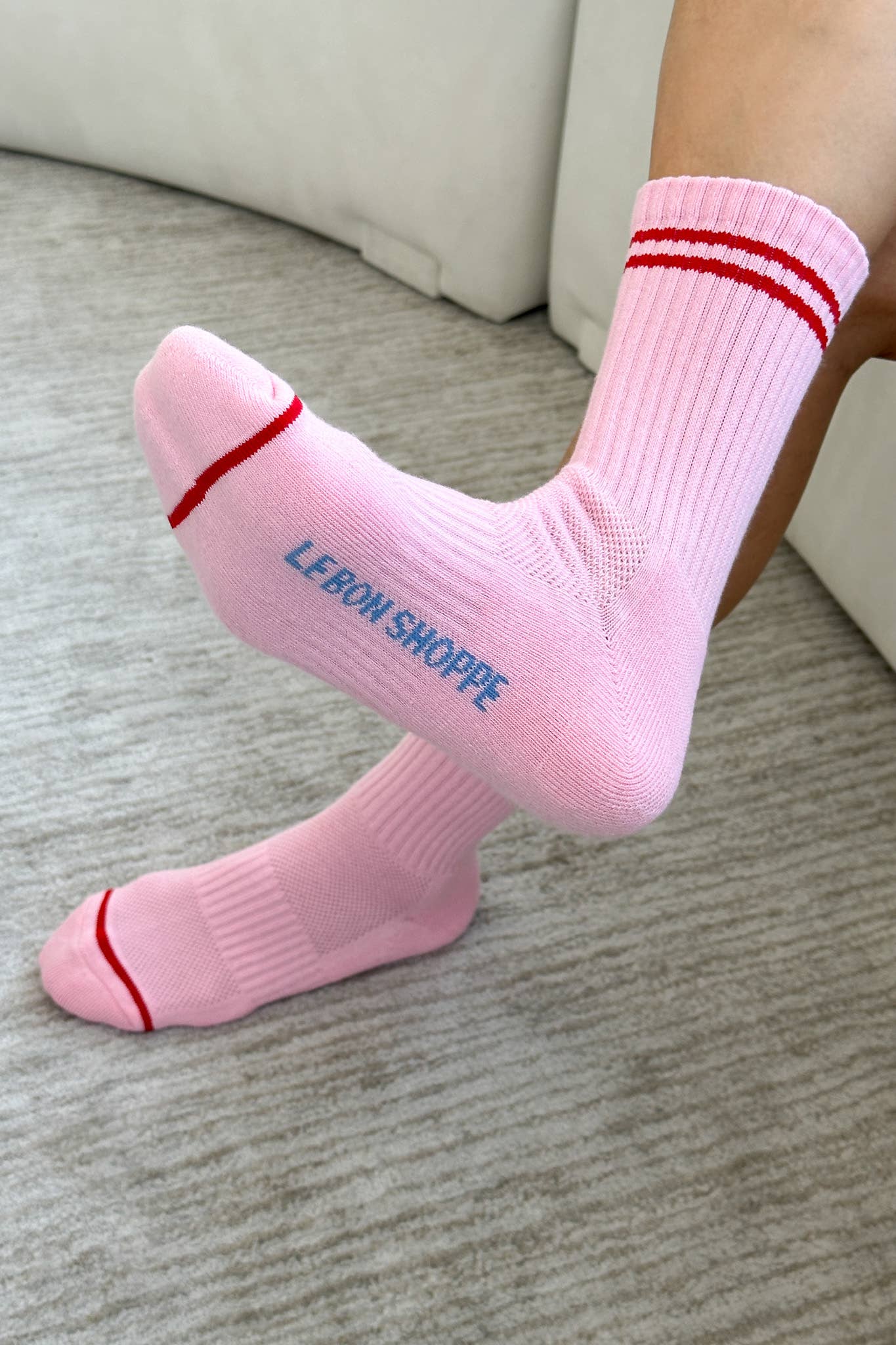 A pair of Le bon shoppe amour pink boyfriend socks which are pink in colour with red stripes at the top and near the toes are worn by a person. It says Le Bon shoppe on the sole of the socks in blue colour. The floor is wooden and is beige with brown lines of detail. 