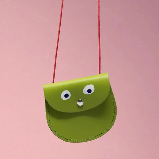 An oval shaped apple green small pocket purse with red thin rope as the bag strap hangs against a pink background. Printed onto the bag are two white googly eyes with black pupils and a small silver press button which acts as the nose and opens/closes the purse. 