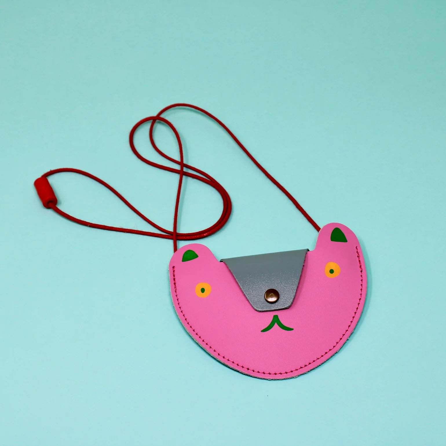 A hot pink pocket purse in the shape of a bears head lays flat on a teal background. The flap of the bag is silver and acts as the nose of the bear and has a black button. The eyes are orange circles with a black pupil. The ears are green. Beneath the flap the mouth is also green. The strap of the bag is thin red rope .