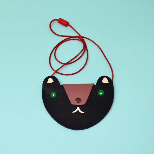 A navy pocket purse in the shape of a bears head laying on a teal background. The flap of the bag is a dusty pink that acts as the nose of the bear and has a a bronze button. The eyes are green circles with a white pupil and the ears are cream. Beneath the flap the mouth is also cream. The strap of the bag is thin red rope. 