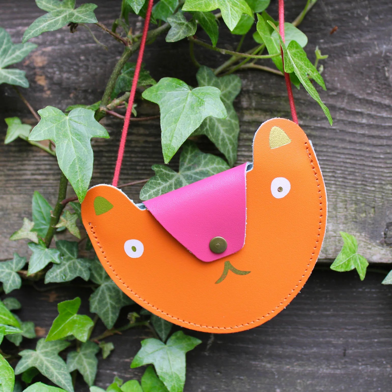 An orange pocket purse in the shape of a bears head hanging in front of what appears to be a wooden fence with leaves surrounding it. The flap of the bag is a ho pink that acts as the nose of the bear and has a a bronze button. The eyes are white circles with gold metallic pupils and the ears are also metallic gold. Beneath the flap the mouth is also metallic gold. The strap of the bag is thin red rope .  