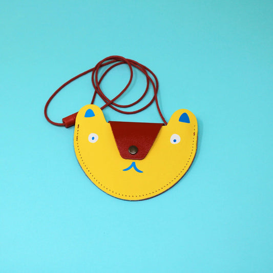 A yellow pocket purse in the shape of a bears head laying flat on a teal background. The flap of the bag is a metallic red that acts as the nose of the bear and has a bronze button. The eyes are white circles with blue pupils and the ears are also blue. Beneath the flap the mouth is also blue. The strap of the bag is thin red rope . 