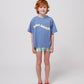 A young child with curly red hair and freckles wears a relaxed-fit blue Bobo Choses Wavy T-Shirt with a cream wavy logo across the chest. They pair it with patterned green and white shorts and bright red jelly sandals. The child has a gentle smile and stands confidently against a plain white background.