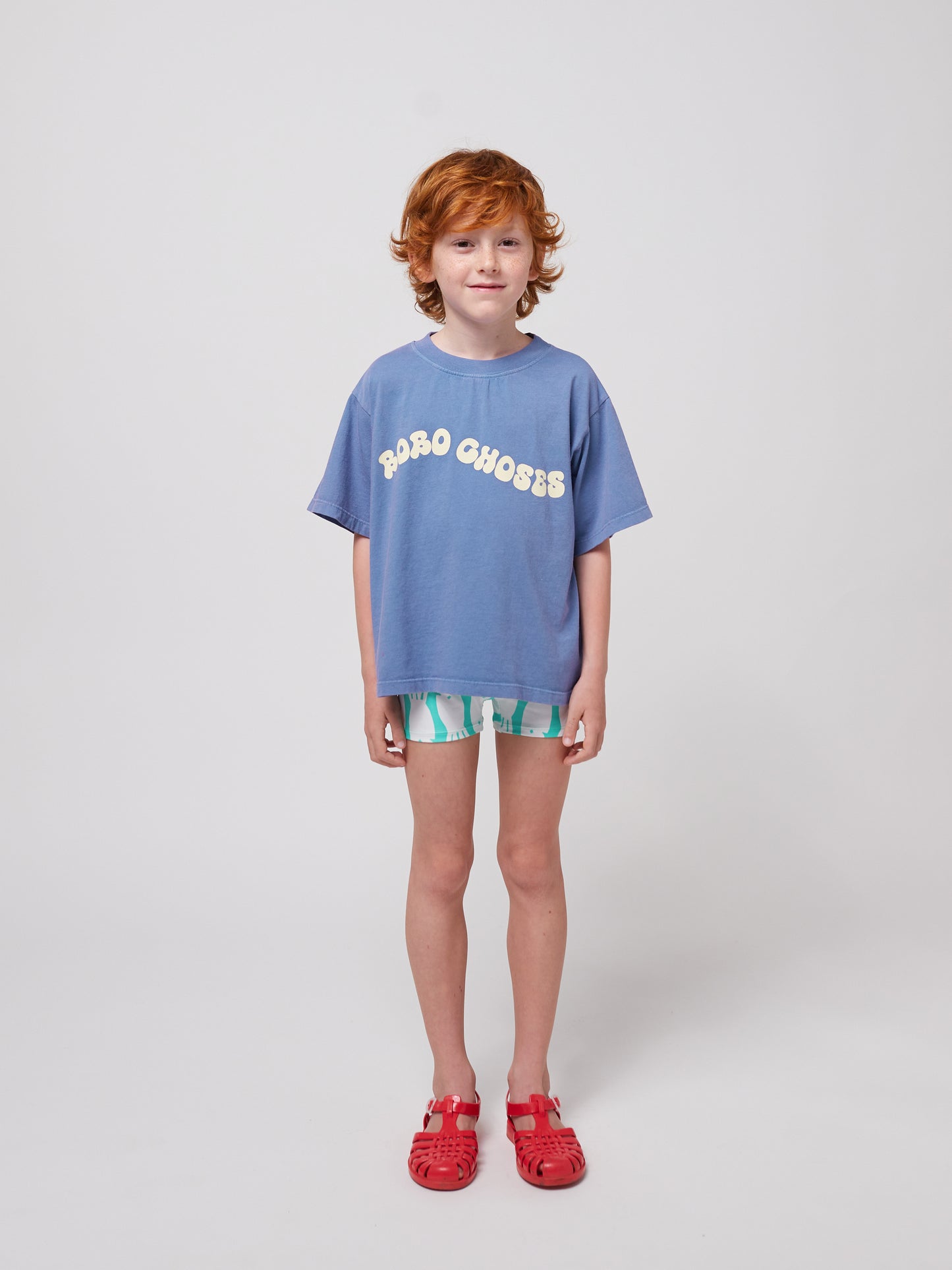 A young child with curly red hair and freckles wears a relaxed-fit blue Bobo Choses Wavy T-Shirt with a cream wavy logo across the chest. They pair it with patterned green and white shorts and bright red jelly sandals. The child has a gentle smile and stands confidently against a plain white background.