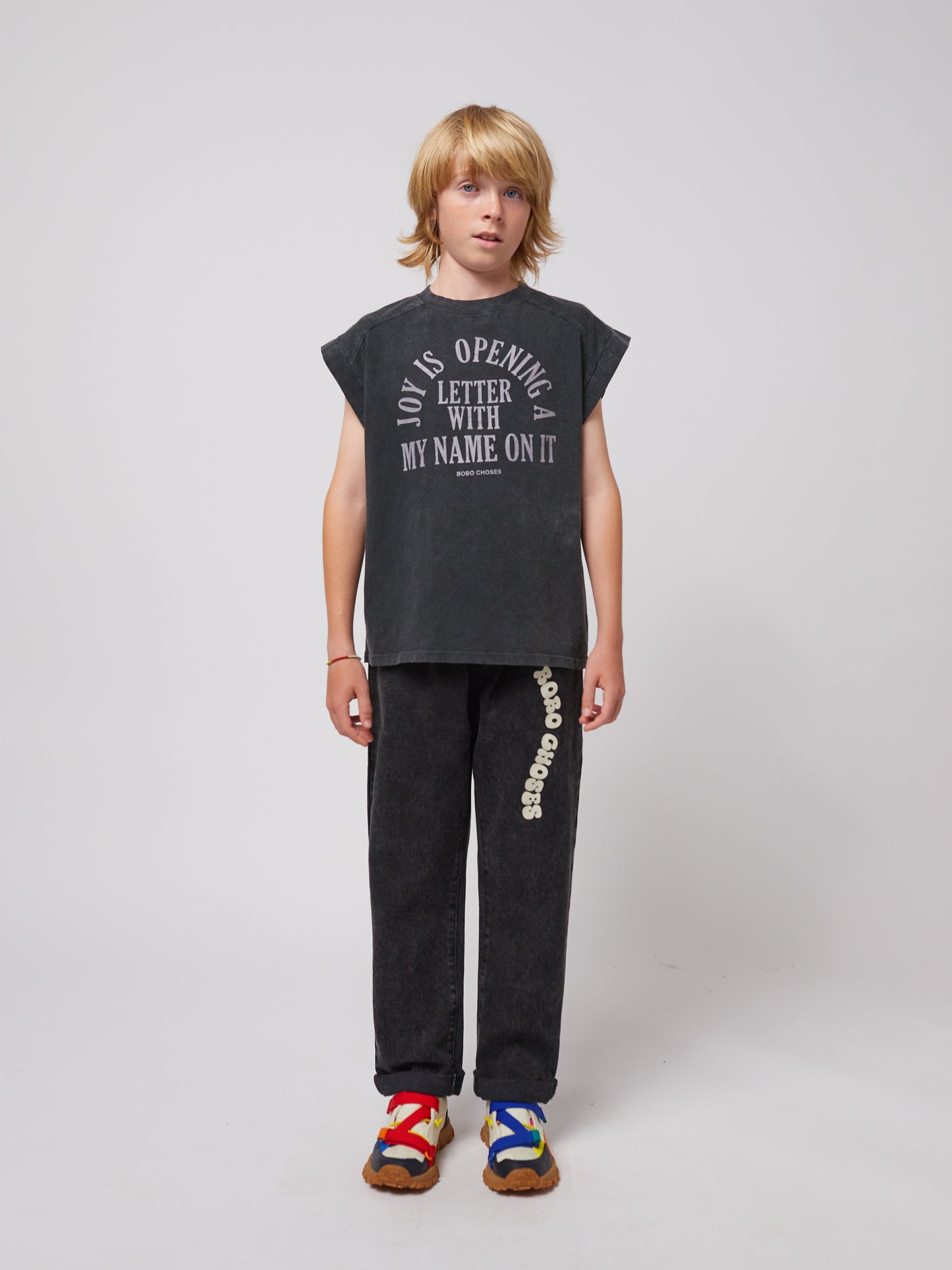 A young model wears the trousers with a black sleeveless graphic t-shirt featuring the text "Joy is opening a letter with my name on it." The trousers have a relaxed fit and are paired with multicoloured sporty sandals.