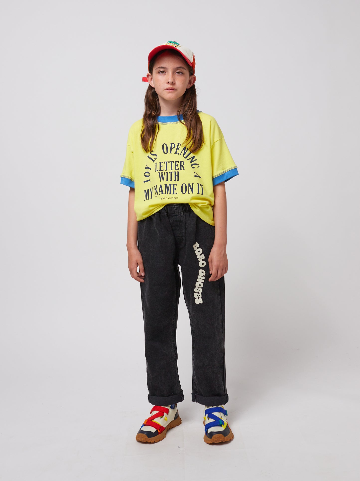 A young model wears the trousers paired with a yellow graphic t-shirt, colourful sneakers, and a red-and-white cap. The relaxed fit and casual style of the trousers are showcased.