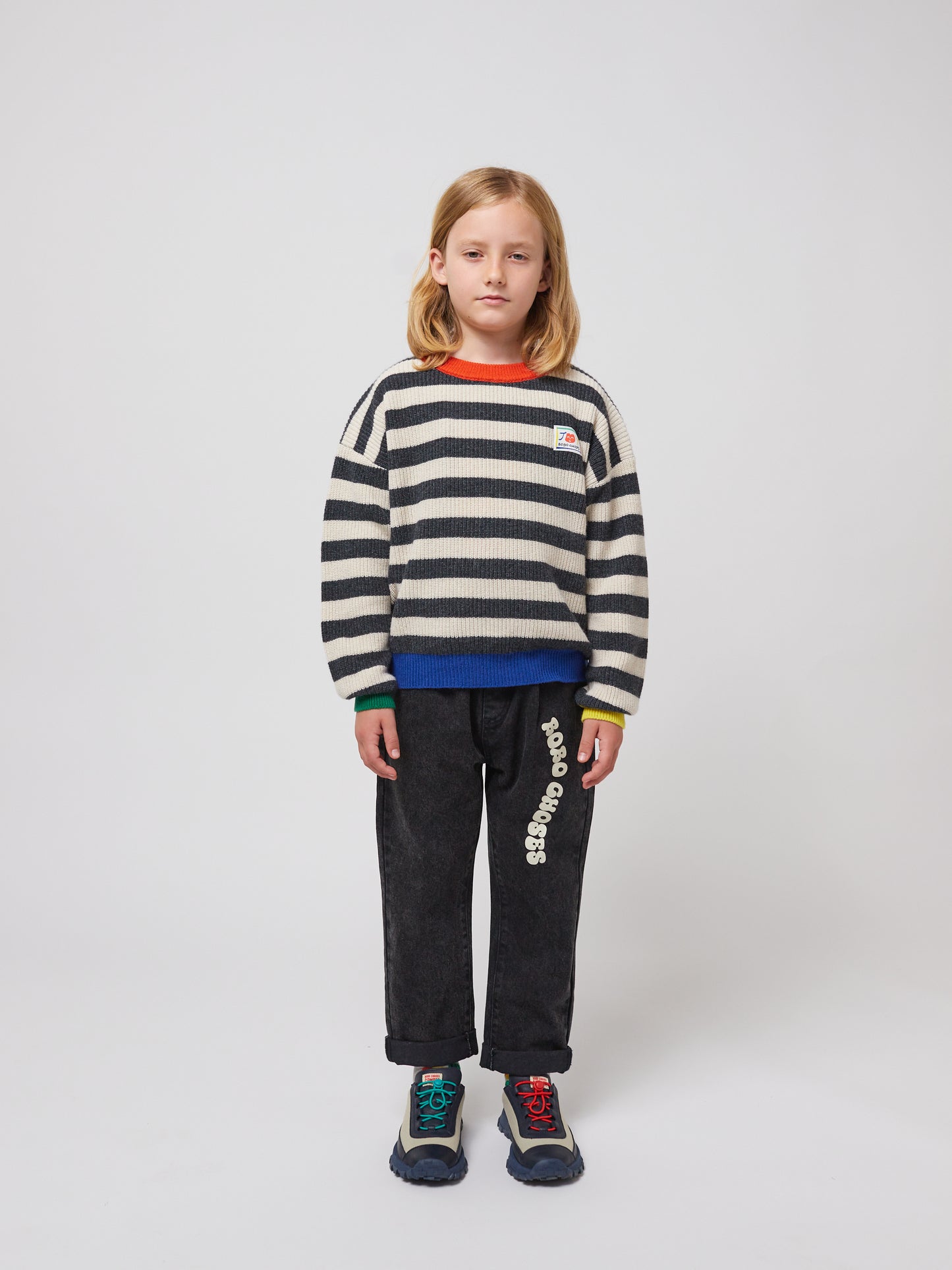 Another child models the trousers with a black-and-white striped knitted jumper, blue-and-black trainers, and the trousers slightly cuffed at the hem.