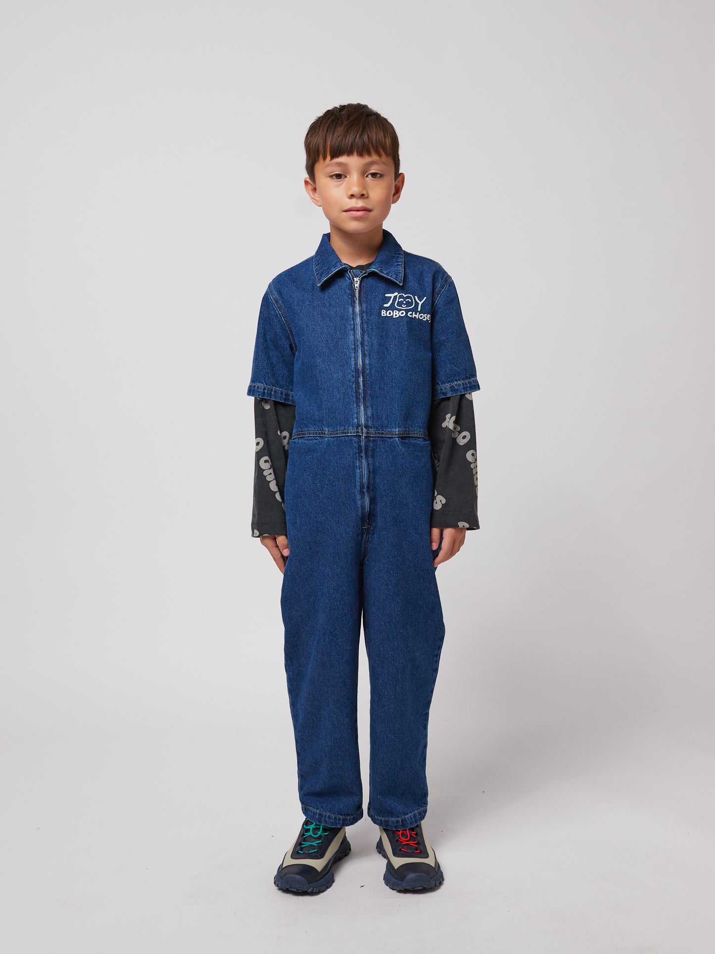 A young child with short dark hair stands facing forward, wearing a dark blue denim jumpsuit with short sleeves, a structured collar, and a front zip closure. The "JOY BOBO CHOSES" logo with a smiling face is printed in white on the left chest. A long-sleeved black top with white "BOBO CHOSES" lettering peeks out from under the jumpsuit. The child is also wearing outdoor-style trainers with mismatched laces – one blue and one red. The background is plain white.