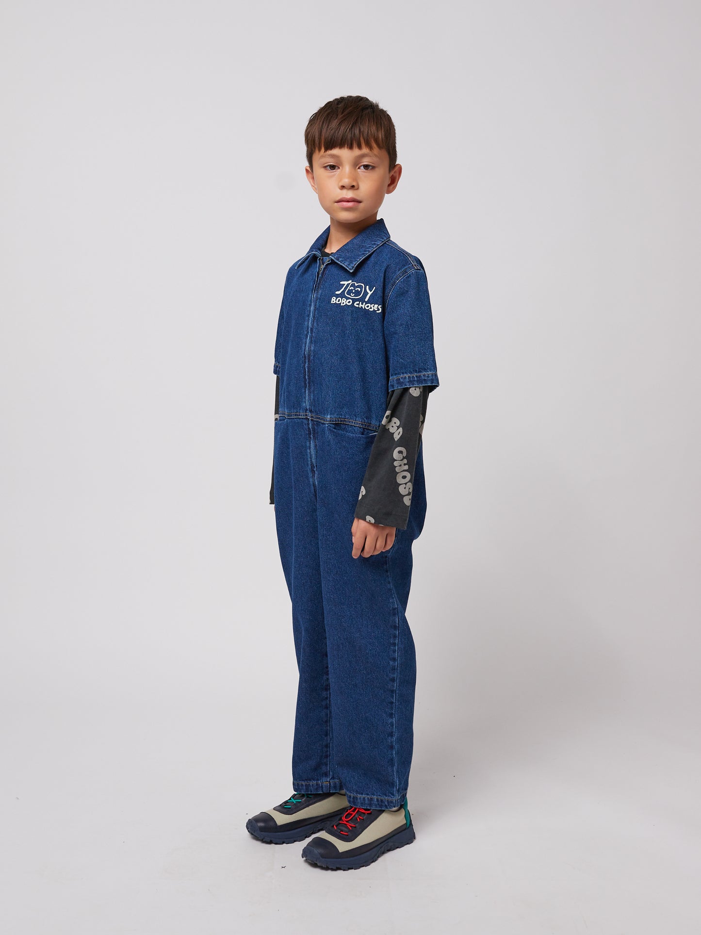 A young child with short dark hair stands slightly turned to the side, wearing a dark blue denim jumpsuit with short sleeves, a structured collar, and a front zip closure. The "JOY BOBO CHOSES" logo with a smiling face is printed in white on the left chest. A long-sleeved black top with white "BOBO CHOSES" lettering extends past the jumpsuit sleeves. The child is also wearing outdoor-style trainers with mismatched laces – one blue and one red. The background is plain white.