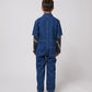 A young child with short dark hair is seen from the back, wearing a dark blue denim jumpsuit with short sleeves and a relaxed fit. The jumpsuit features visible stitching details, a structured collar, and a single square patch pocket on the right side of the lower back. A long-sleeved black top with white "BOBO CHOSES" lettering extends past the jumpsuit sleeves. The child is also wearing outdoor-style trainers with mismatched laces – one blue and one red. The background is plain white.