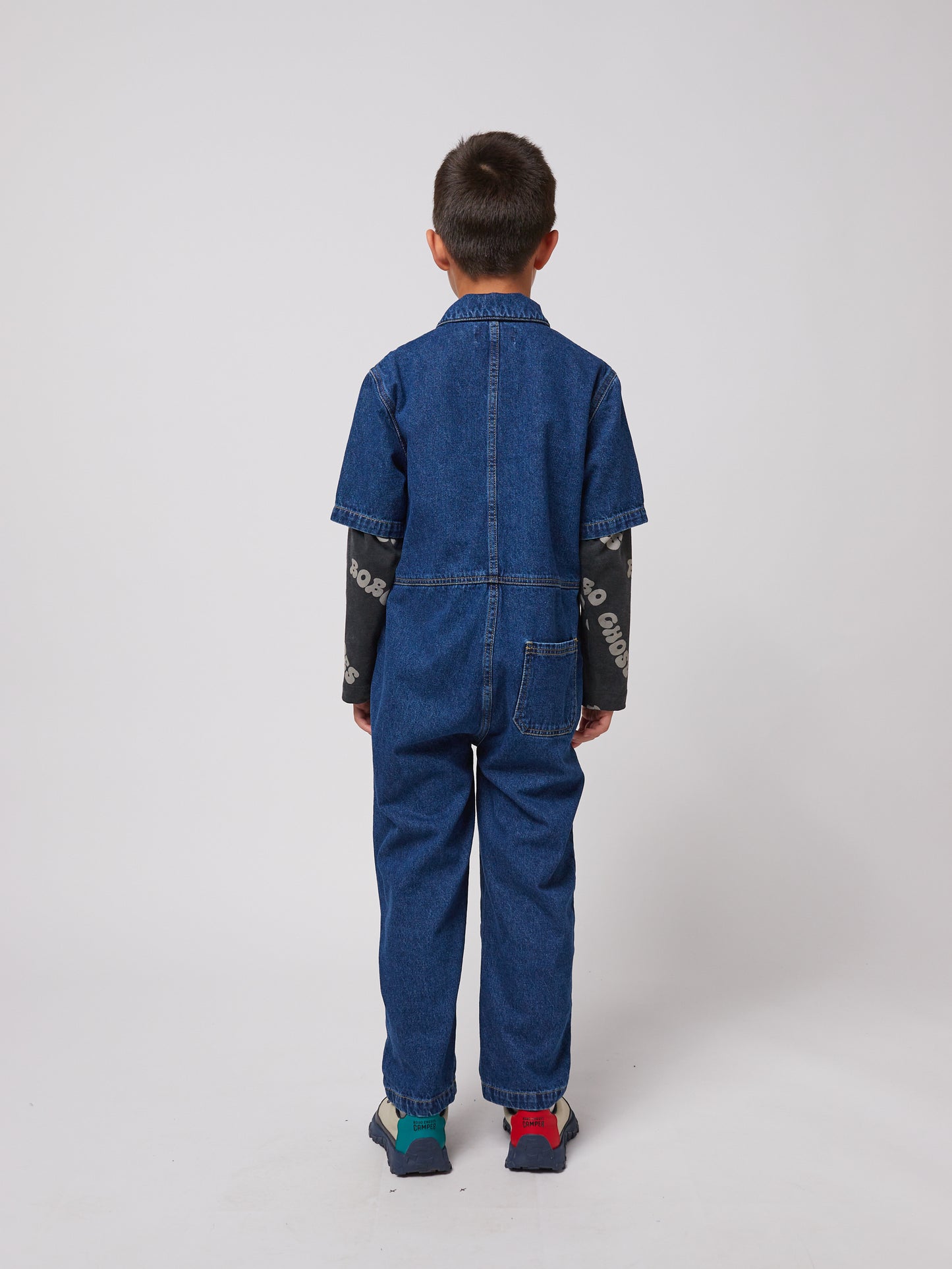 A young child with short dark hair is seen from the back, wearing a dark blue denim jumpsuit with short sleeves and a relaxed fit. The jumpsuit features visible stitching details, a structured collar, and a single square patch pocket on the right side of the lower back. A long-sleeved black top with white "BOBO CHOSES" lettering extends past the jumpsuit sleeves. The child is also wearing outdoor-style trainers with mismatched laces – one blue and one red. The background is plain white.